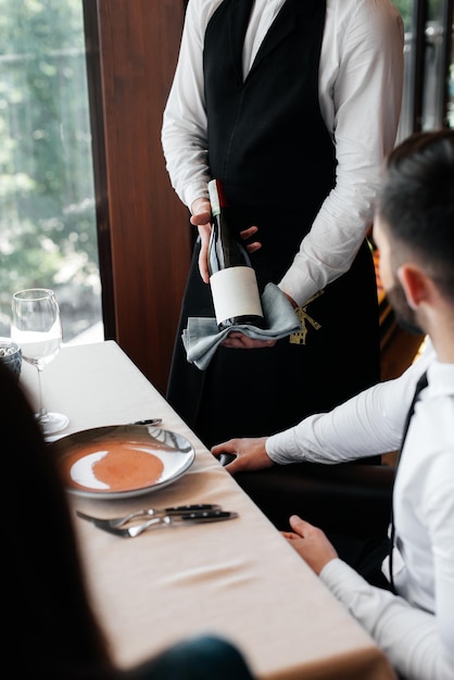 세련된 앞치마를 입은 젊은 소믈리에가 레스토랑에서 아름다운 커플에게 고급 와인을 시연하고 제공합니다. 엘리트 레스토랑과 공공 레스토랑에서 고객 서비스 프리미엄 사진
