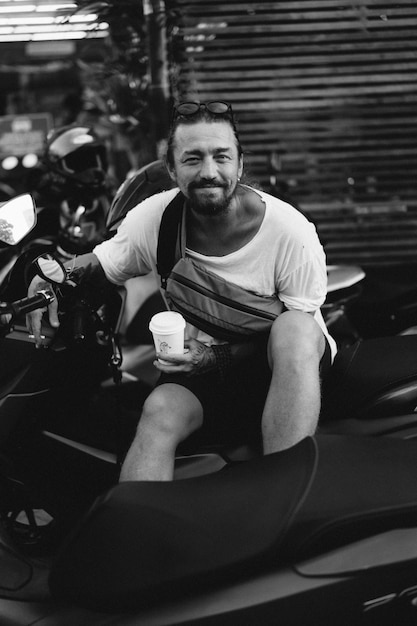 あごひげを生やした若いスタイリッシュな男は、使い捨ての紙コップからコーヒーを飲み、通りのバイクに座っています。ライフスタイル、トロピカルライフ。バリ