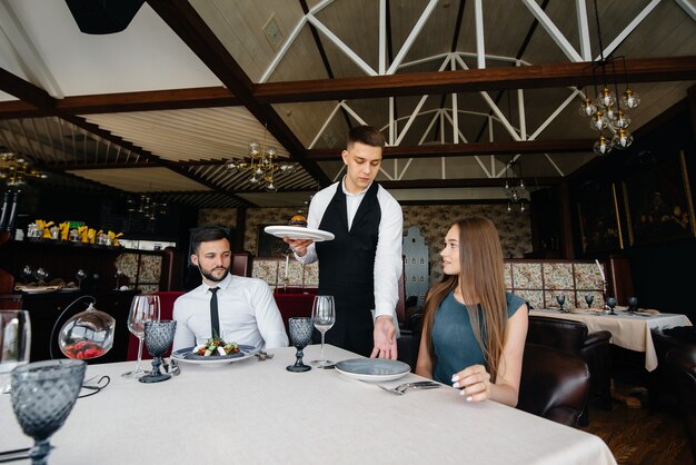 세련된 앞치마를 입은 젊은 웨이터가 세련된 레스토랑에서 아름다운 부부와 함께 테이블을 제공합니다. 고객 서비스.