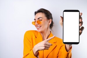 a young woman in trendy stylish glasses and a bright orange oversized jacket on a white background holds a phone with a blank white screen
