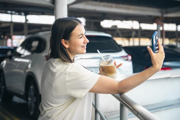 無料写真 駐車場でコーヒーを飲みながら自撮りする若い女性