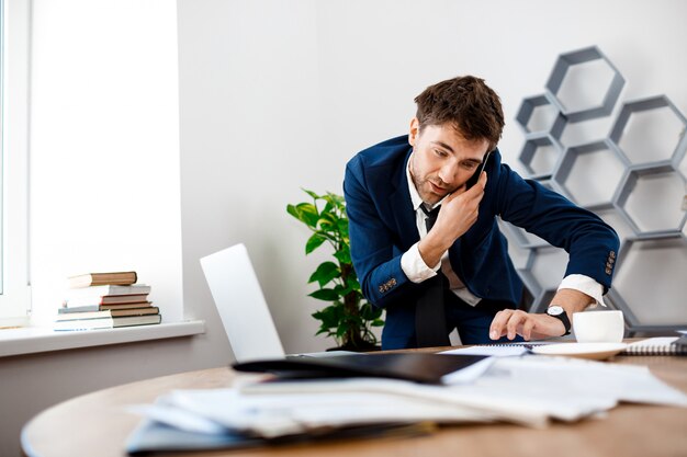 Absentminded young businessman speaking on phone, office background.