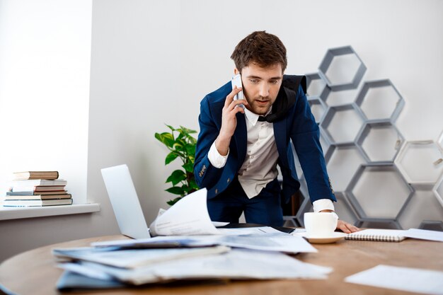 Absentminded young businessman speaking on phone, office background.