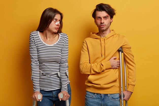 Free photo accident woman and man walk with crutches, have problems with health after dangerous ride on motorbike, being reckless drivers, come to doctor for consultation, stand indoor over yellow wall