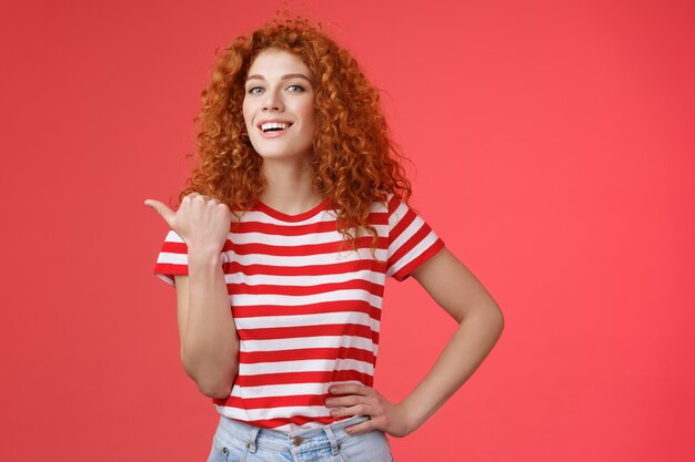 Accomplished good-looking redhead curly woman raise head proudly confident pointing thumb left smiling camera assertive sharing awesome product location recommend perfect store offer.