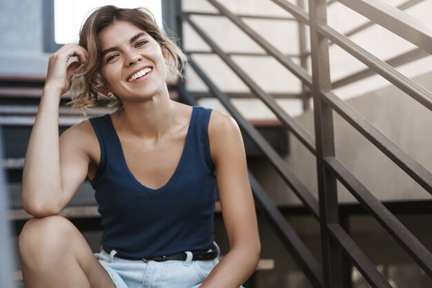 Accomplished happy carefree young blond european girl in blue top, shorts sit relaxed outdoor modern stairs tilting head cute, smiling toothy having fun enjoying good day, talking upbeat.