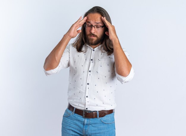 Aching adult handsome man wearing glasses keeping hands on head with closed eyes isolated on white wall