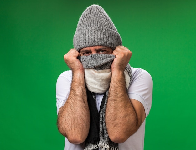 Free photo aching adult ill caucasian man covering face with scarf and wearing winter hat isolated on green wall with copy space