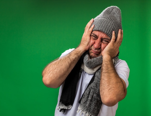 aching adult ill caucasian man with scarf around neck wearing winter hat holding head isolated on green wall with copy space