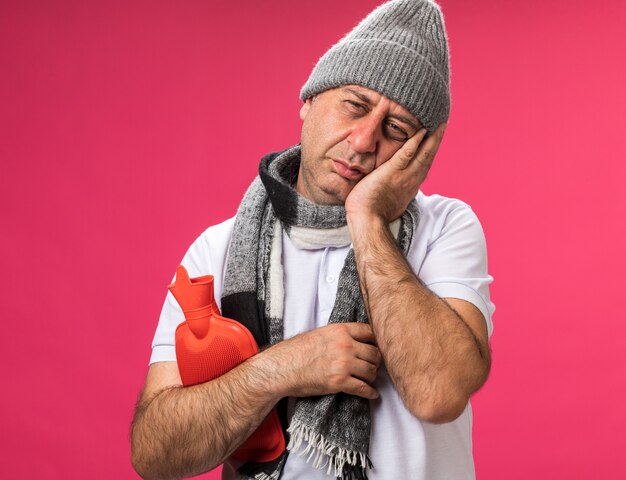 aching adult ill caucasian man with scarf around neck wearing winter hat puts hand on face and holds hot water bottle isolated on pink wall with copy space