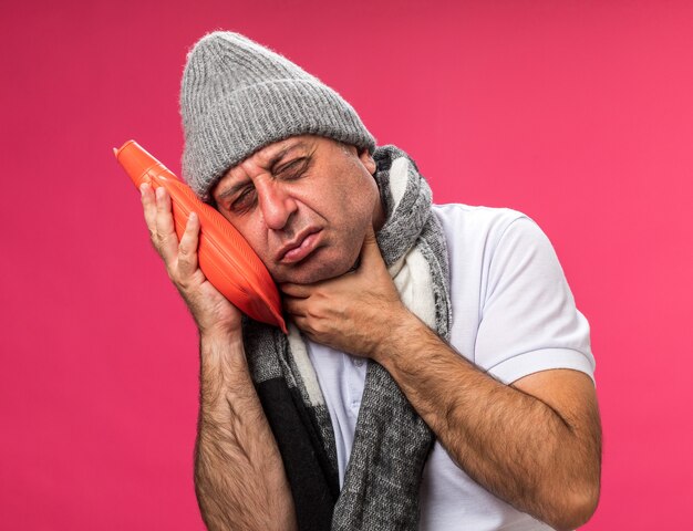 aching adult ill caucasian man with scarf around neck wearing winter hat puts hand on neck and holds hot water bottle isolated on pink wall with copy space