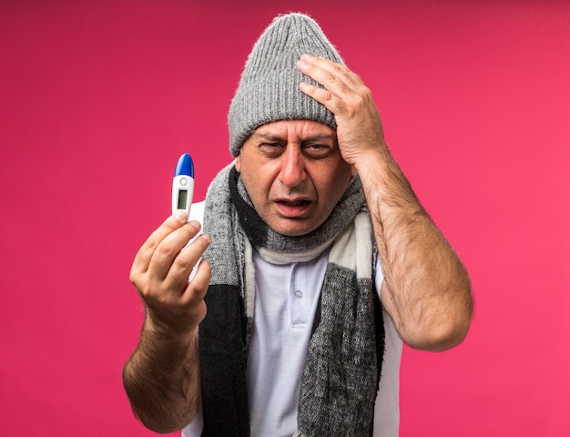 aching adult ill caucasian man with scarf around neck wearing winter hat putting hand on head and holding thermometer isolated on pink wall with copy space