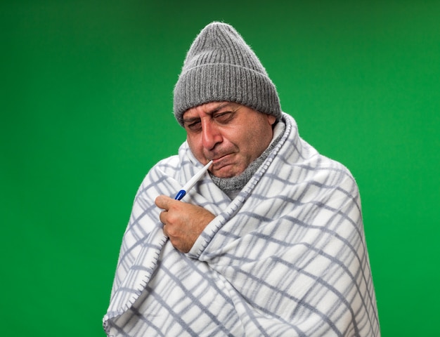 больной взрослый больной кавказский мужчина с шарфом на шее в зимней шапке, завернутый в плед, держит во рту термометр, изолированный на зеленой стене с копией пространства