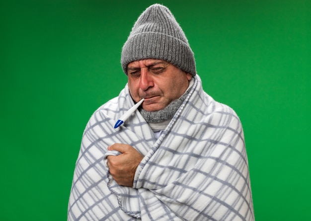 Uomo caucasico malato adulto dolorante con sciarpa intorno al collo che indossa cappello invernale avvolto in supporti a quadri con gli occhi chiusi tenendo il termometro in bocca isolato sul muro verde con spazio di copia