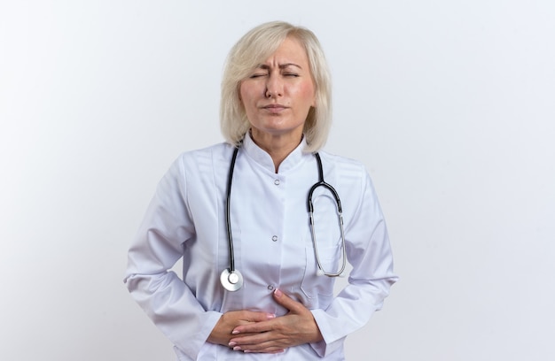 Foto gratuita medico femminile slava adulto dolorante in abito medico con stetoscopio che tiene la sua pancia isolata su fondo bianco con lo spazio