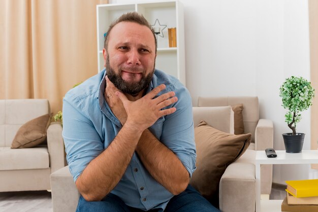 Больной взрослый славянский мужчина сидит на кресле, держась за шею двумя руками в гостиной