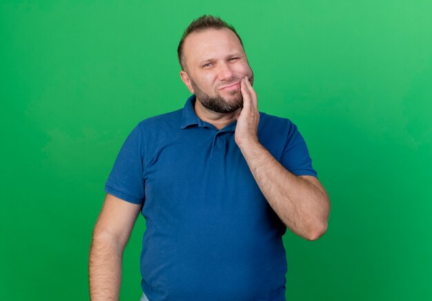 Больной взрослый славянский мужчина трогает щеку, страдающую от зубной боли, изолированную на зеленой стене с копией пространства