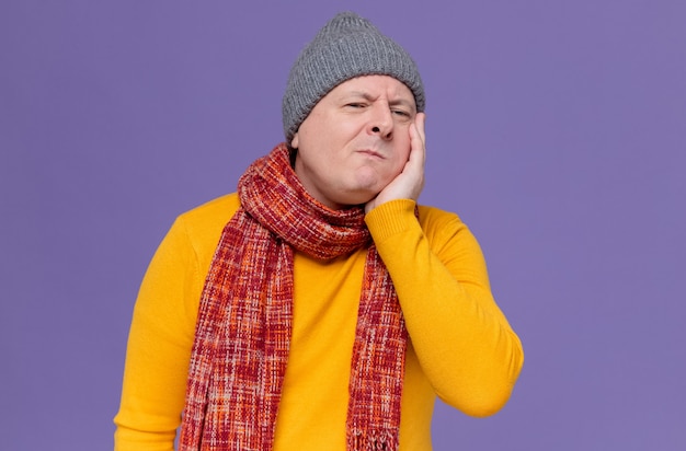 Aching adult slavic man with winter hat and scarf around his neck putting hand on his face