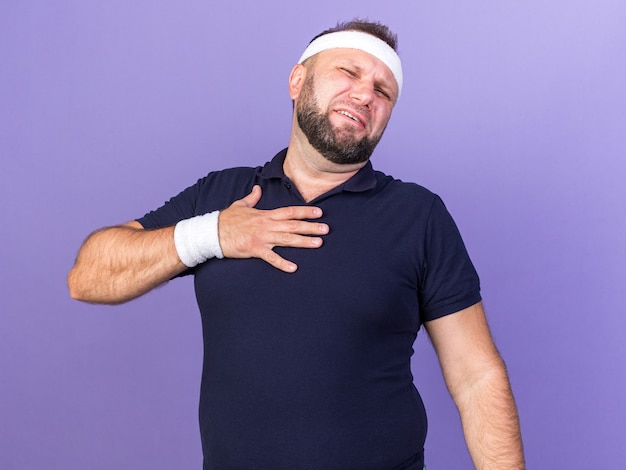 больной взрослый славянский спортивный мужчина с повязкой на голову и браслетами, положив руку на грудь, изолированную на фиолетовой стене с копией пространства
