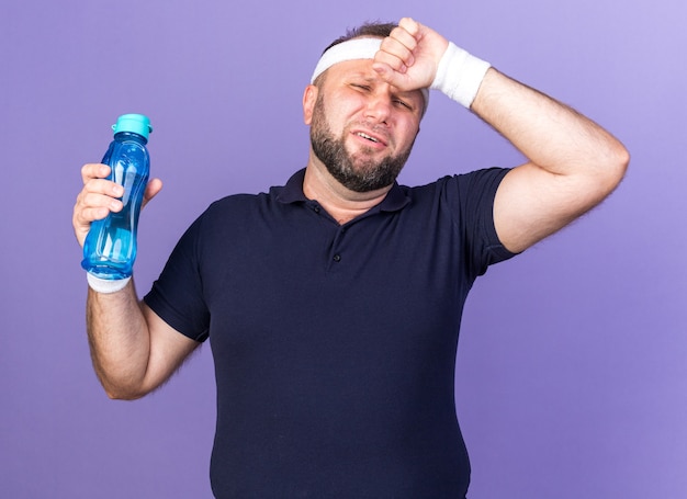 Dolorante adulto slavo sportivo uomo che indossa la fascia e braccialetti mettendo la mano sulla fronte e tenendo la bottiglia di acqua isolata sul muro viola con spazio di copia