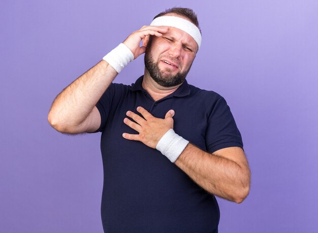 больной взрослый славянский спортивный мужчина с повязкой на голову и браслетами, положив руку на голову и грудь, изолированные на фиолетовой стене с копией пространства