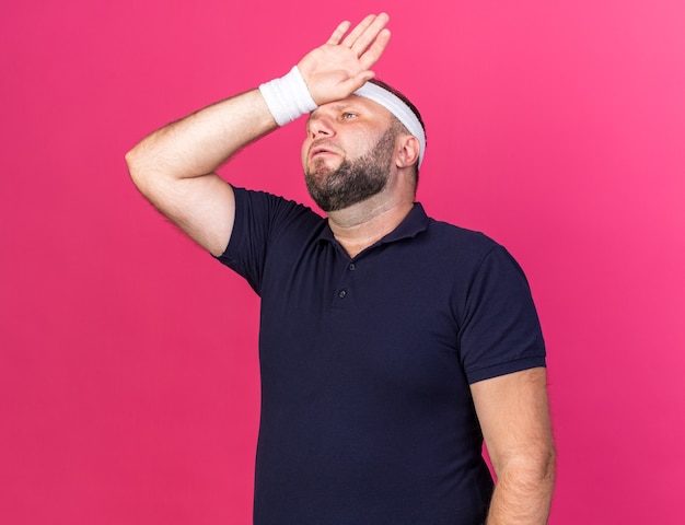 больной взрослый славянский спортивный мужчина с повязкой на голову и браслетами, положив руку на лоб, изолированный на розовой стене с копией пространства