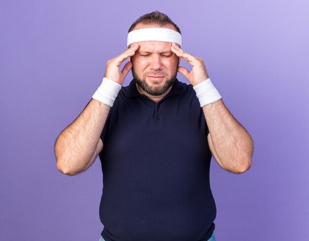 aching adult slavic sporty man wearing headband and wristbands putting hands on forehead isolated on purple wall with copy space