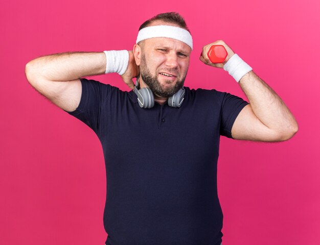 больной взрослый славянский спортивный мужчина в наушниках с повязкой на голову и браслетами держит гантель и кладет руку ему на шею, изолированную на розовой стене с копией пространства