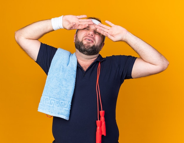 aching adult slavic sporty man with jumping rope around neck wearing headband and wristbands holding towel on shoulder and putting hands on forehead isolated on orange wall with copy space