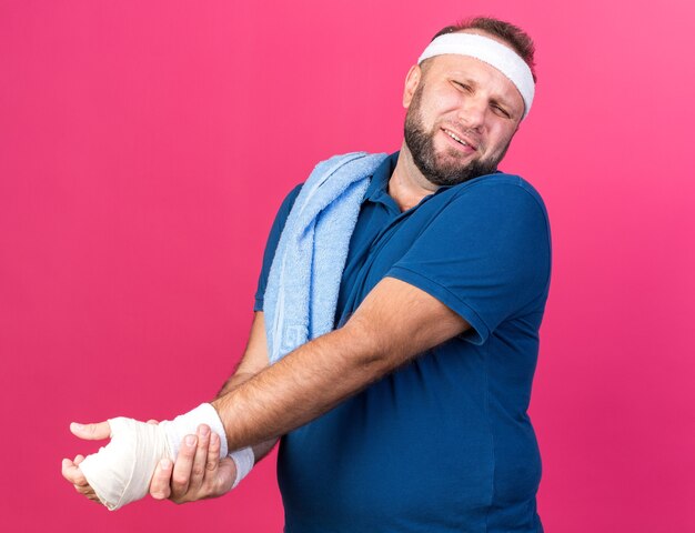 Бесплатное фото Больной взрослый славянский спортивный мужчина с полотенцем на плече, носящий повязку на голову и браслеты, держащий руку, изолированную на розовой стене с копией пространства