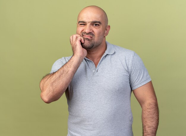 Aching casual middle-aged man  keeping hand on cheek suffering from toothache isolated on olive green wall