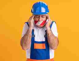 Free photo aching caucasian adult builder man in uniform covered mouth with duct tape puts hands on temples on orange