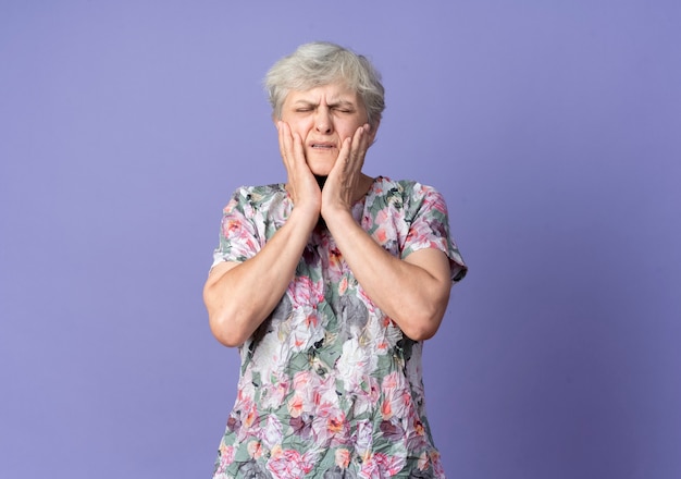 Бесплатное фото Больная пожилая женщина кладет руки на лицо, изолированное на фиолетовой стене