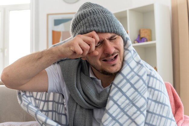 aching ill man with scarf around neck wearing winter hat wrapped in plaid putting his hand on head sitting with closed eyes on couch at living room