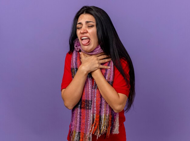 Aching and irritated young caucasian ill girl wearing scarf holding throat with closed eyes isolated on purple wall with copy space