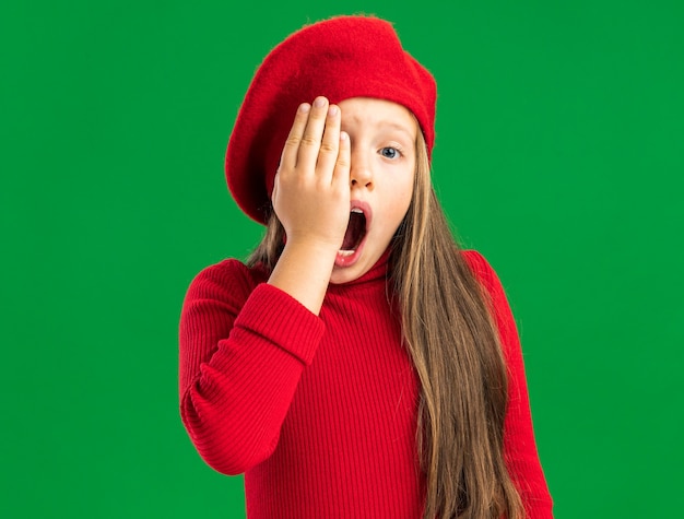 Aching little blonde girl wearing red beret keeping hand on eye with open mouth isolated on green wall with copy space