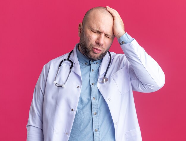 Aching middle-aged male doctor wearing medical robe and stethoscope keeping hand on head suffering from headache with closed eyes isolated on pink wall