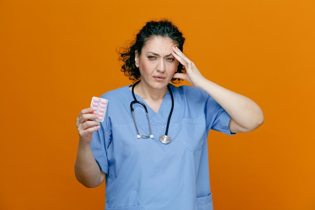 Больная женщина-врач средних лет в униформе и со стетоскопом на шее показывает упаковку капсул, смотрит в камеру, держа руку на голове, с головной болью на оранжевом фоне