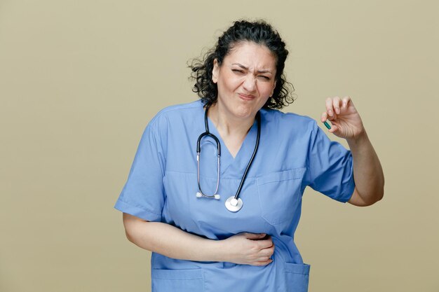 Aching middleaged female doctor wearing uniform and stethoscope around neck showing capsule looking at camera keeping hand on belly isolated on olive background