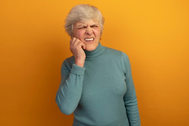 Aching old woman wearing blue turtleneck sweater looking at side touching cheek suffering from toothache isolated on orange wall with copy space
