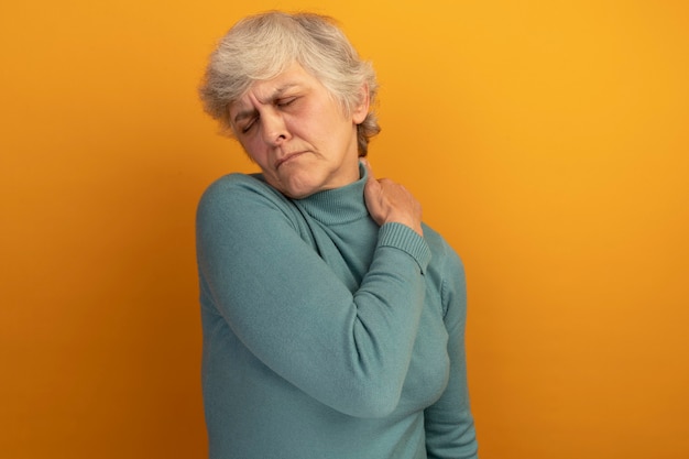 Free photo aching old woman wearing blue turtleneck sweater putting hand on shoulder with closed eyes