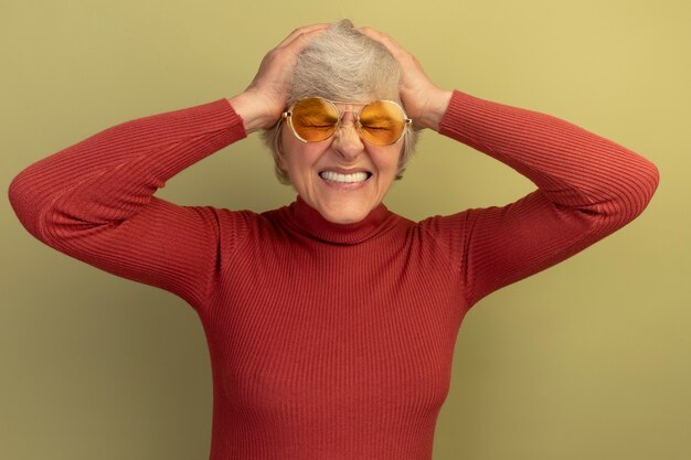 Aching old woman wearing red turtleneck sweater and sunglasses keeping hands on head suffering from headache with closed eyes isolated on olive green wall