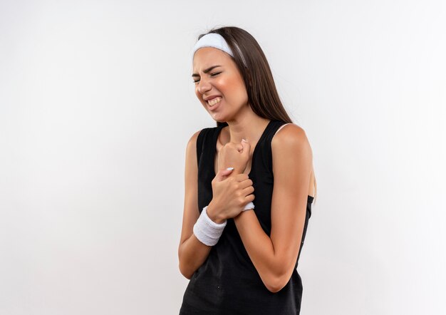 Aching pretty sporty girl wearing headband and wristband holding arm with closed eyes isolated on white wall with copy space