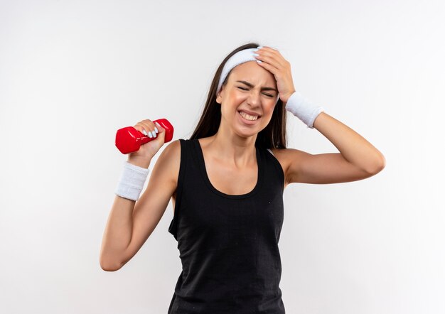 Aching pretty sporty girl wearing headband and wristband holding dumbbell suffering from headache with hand on head and closed eyes isolated on white wall