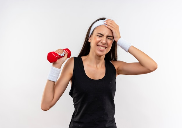 Aching pretty sporty girl wearing headband and wristband holding dumbbell suffering from headache with hand on head and closed eyes isolated on white wall