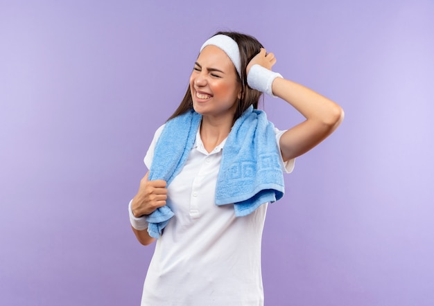 Aching pretty sporty girl wearing headband and wristband putting hand on head closing eyes with towel around neck isolated on purple wall