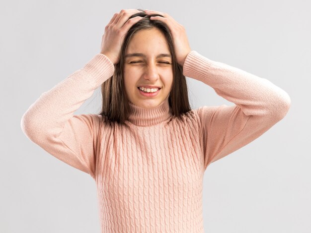 Aching pretty teenage girl keeping hands on head with eyes closed showing tooths isolated on white wall