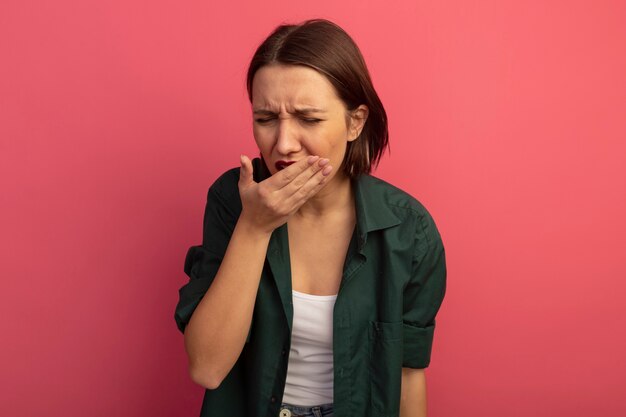 Aching pretty woman puts hand on mouth isolated on pink wall