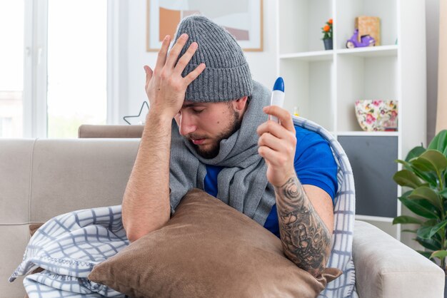 スカーフと冬の帽子を身に着けている痛くて弱い若い病気の人は、目を閉じて頭に手を保ちながら温度計を保持している彼の足に枕を置いてリビングルームのソファに座っています