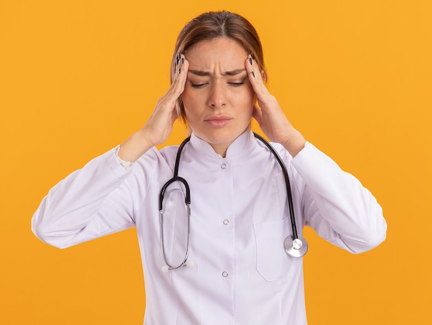 Aching with closed eyes young female doctor wearing medical robe with stethoscope putting hands on temple isolated on yellow wall